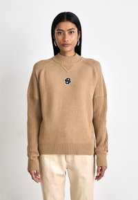 Suéter de malha bege com mangas e bainha caneladas, apresentando um logotipo preto no peito. Textura suave, corte casual, gola alta.