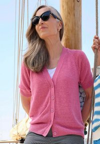 Hellrosa Strickjacke mit kurzen Ärmeln, Knopfverschluss und leicht strukturiertem Strickmuster. Über einem weißen Oberteil mit schwarz-weißen karierten Hosen getragen.