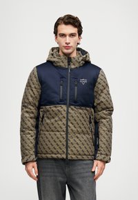 Homemem vestido com uma jaqueta capuz azul-marinho e marrom com padrão, com bolsos com zíper, de pé contra um fundo branco liso.