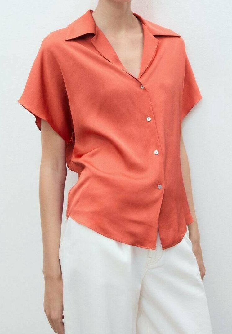 Femme portant un chemisier corail à manches courtes amples avec col et boutons, associé à un pantalon blanc taille haute.