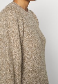 Beige gestrickter Pullover mit lockerer Passform, der ein strukturiertes Stichmuster und einen runden Ausschnitt aufweist und weiche Fasern sowie minimale Nähte zeigt.