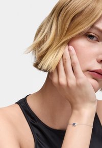 Pulsera de plata con una piedra azul, llevada en una muñeca. El portador tiene el pelo corto y rubio y reposa su mentón sobre su mano, vistiendo una blusa negra.