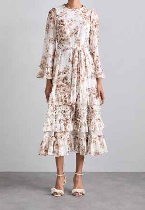 Florales Midikleid in Weiß mit braunen Tönen, gestuften Rüschen, langen Ärmeln und einem Taillengürtel. Kombiniert mit hellfarbenen Sandaletten mit Absatz.