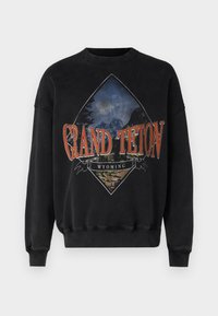 Felpa nera a collo rotondo con una grafica di un paesaggio montano e il grande testo arancione "Grand Teton Wyoming" sul davanti.