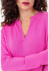 Pull en maille rose avec un col en V. Les manches longues ont une coupe décontractée. La texture semble douce et légère.