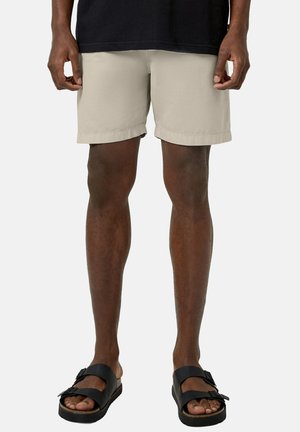 Person, die beige Shorts und schwarze Sandalen mit doppeltem Riemen trägt, steht mit entspannten Händen an den Seiten vor schlichtem Hintergrund.