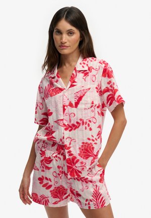 Femme portant une chemise blanche à manches courtes et un short assortis avec des imprimés floraux rouges, debout avec une main dans la poche.