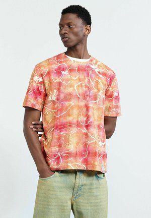 Muškarac obučen u crvenu i narančastu košulju kratkih rukava s cvjetnim uzorkom i svijetloplave traperice, gleda u stranu s jednom rukom u džepu.