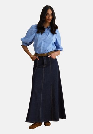 Frau trägt eine blau gestreifte Bluse mit Puffärmeln, einen braunen Gürtel, einen langen dunklen Denimrock und braune Schuhe, steht mit den Händen in den Taschen.