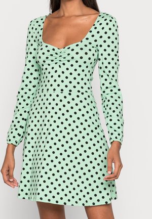 Robe vert menthe à manches longues, à pois noirs, avec un décolleté carré et un détail froncé à l'avant. Tissu doux avec une jupe évasée.