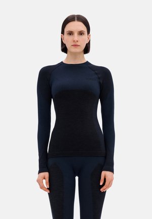 Frau mit kurzem dunklem Haar, die ein eng anliegendes langärmliges schwarzes und marineblaues Sportoberteil sowie passende Leggings trägt und vor einem schlichten Hintergrund steht.