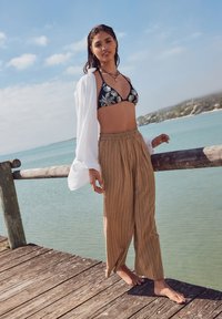 Top bikini floreale blu abbinato a pantaloni larghi a righe beige. Un copricostume bianco trasparente completa l'outfit, su un pontile in legno vicino all'acqua.