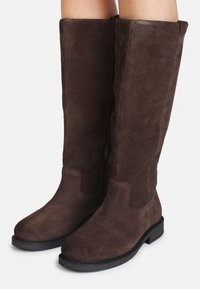 Bottes en suedine marron, montantes jusqu'au genou, avec une texture lisse, un petit talon noir et une fermeture éclair sur le côté. Elles présentent un design à bout arrondi.