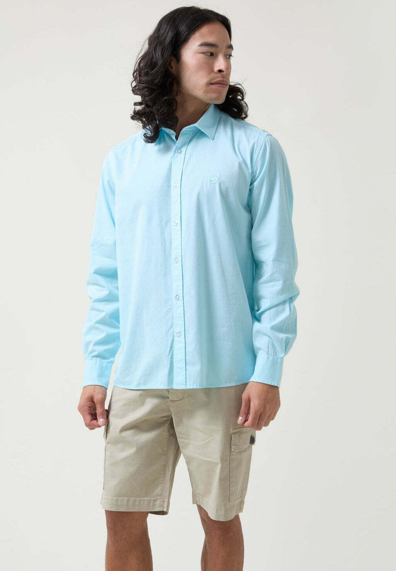 Camisa de botonada de manga larga en azul claro con cuello, combinada con pantalones cortos cargo beige que presentan bolsillos laterales y detalles cosidos.