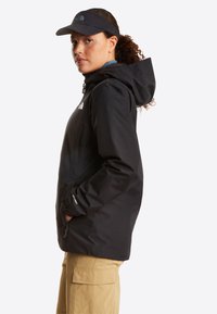 Γυναίκα με μαύρο μπουφάν με κουκούλα και μαύρο καπέλο με γείσο, κοιτάζοντας πλάγια, στέκεται με τα χέρια στις τσέπες του μπουφάν μπροστά σε λευκό φόντο.