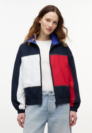 Junge Frau, die eine marineblaue, weiße und rote Jacke mit Reißverschluss und blauem Kragen über einem weißen Hemd und hellblauen Jeans trägt, stehend vor einem einfarbigen Hintergrund.