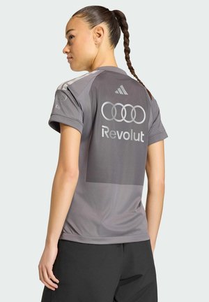 Donna con capelli intrecciati che indossa una maglietta sportiva grigia Adidas con i loghi Audi e Revolut sul retro, in piedi davanti a uno sfondo neutro.