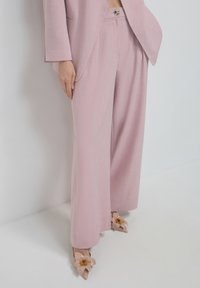 Helle rosa, weit geschnittene Hosen aus strukturiertem Stoff. Kombiniert mit hautfarbenen High Heels, die Blumendetails auf den Zehen haben.