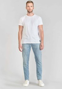 Camiseta de algodón blanca combinada con jeans de mezclilla azul claro. El modelo está de pie con las manos a los lados, vistiendo zapatillas blancas, sobre un fondo neutro.