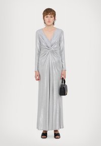 Ασημένιο μεταλλικό μακρύ φόρεμα με μακριά μανίκια, βαθύ V-λαιμόκοψη, λεπτομέρεια στροφής στη μέση και ρέουσα φούστα. Συνδυασμένο με μαύρες γόβες και τσάντα.