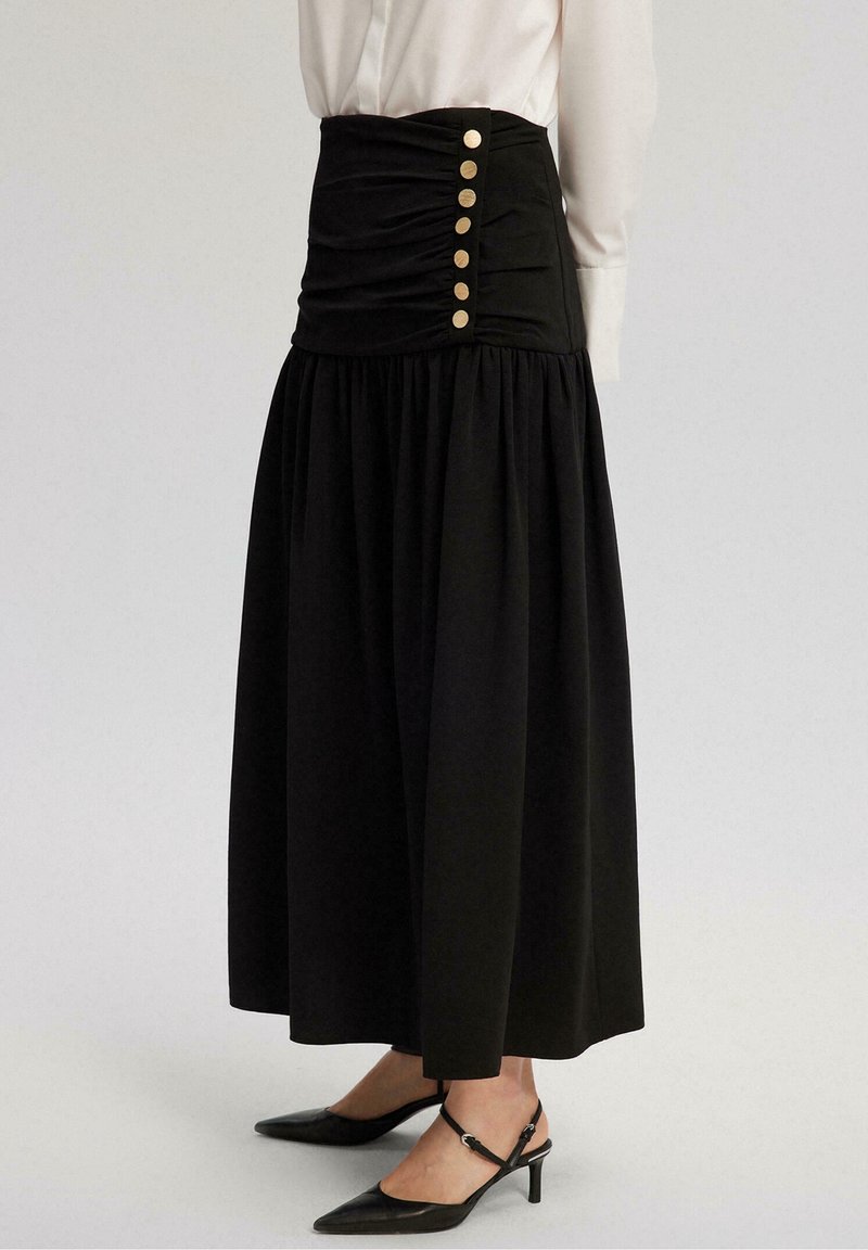 Schwarzer Maxikleid mit gerafftem Bund und goldenen Knopfdetails an der Seite. Kombiniert mit spitzen schwarzen Absätzen und einer weißen Bluse.