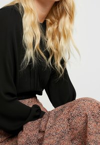 Blouse noire avec des manches bouffantes et des détails plissés, assortie à une jupe à motifs dans des tons orange et rose, avec un tissu doux et texturé.