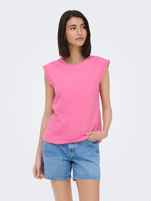 Mujer de pie con una blusa sin mangas rosa con encaje en los bordes y shorts de mezclilla azul, mirando ligeramente hacia un lado con una mano en el bolsillo.