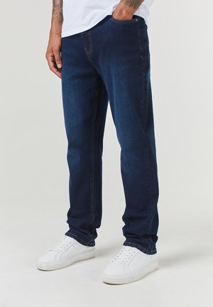 Vaqueros de mezclilla azul oscuro con un ajuste recto, que presentan detalles de costura en naranja y un diseño clásico de bolsillo, combinados con zapatillas blancas.