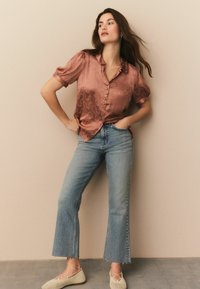 Satin, rosenfarvet skjorte med krave og knaplukning, parret med lyseblå flared jeans. Modellen har beige vævede flade sko på.