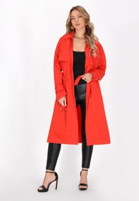 Rode trenchcoat met een ceintuur in de taille, grote kraag en knopdetails. Draagt over een zwarte top en glanzende zwarte legging, met zwarte hoge hakken.