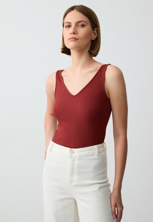 Gerippter bordeauxfarbener Tanktop mit V-Ausschnitt und breiten Tragriemen, kombiniert mit hochgeschnittenen weißen Hosen. Glatte Textur, enganliegendes Design.