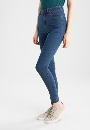 Femme portant un jean skinny taille haute bleu et des baskets beiges, debout devant un fond blanc.
