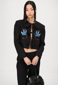 Veste noire courte avec un grand col, ornée d'une broderie d'oiseaux bleus et de accents rouges. Comprend des liens en ruban à l'avant.