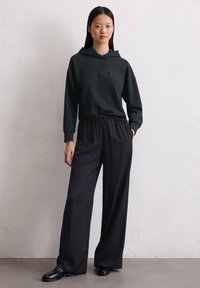 Czarna bluza z kapturem, z wytłoczonym logo marki. Połączona z luźnymi czarnymi spodniami i czarnymi butami, na neutralnym tle.