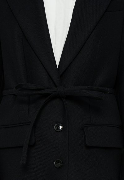 Mustast tekstuuriga kangast jakk, sälguga krae, kaks esist taskut ja vöökohal sõlmitav detail. Omab nööpidega kinnitust.