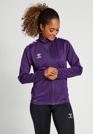 Veste de sport violette avec un col haut, une fermeture éclair intégrale et un tissu texturé. Présente des bandes contrastantes sur les épaules et un logo sur la poitrine.