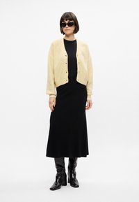 Cardigan en tricot jaune clair à manches longues et boutons, porté par-dessus une robe noire moulante et des bottes noires à talons jusqu'au genou.