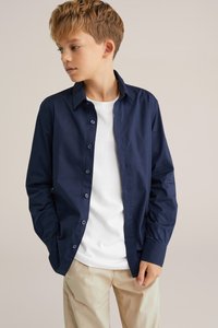 Tamsiai mėlyna marškinė su struktūrine apykakle, ilgomis rankovėmis ir glotnia tekstūra, dėvimi ant balto marškinėlio ir derinami su bežinėmis kelnėmis.