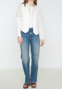 Biała lniana kamizelka na białej koronkowej bluzce z długim rękawem, zestawiona z jasnoniebieskimi dzwonowymi jeansami i brązowymi sandałami na platformie, na białym tle.