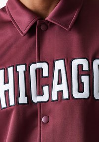 Maroon atletska majica s bijelom oznakom "CHICAGO" s crnim obrubom, glatke teksture i ovratnikom na dugme.