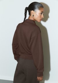 Femme avec une queue de cheval tressée portant des boucles d'oreilles en forme d'étoile, un haut marron à manches longues et un pantalon noir, se tenant devant un mur clair projetant une ombre.