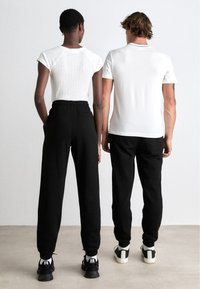 Twee personen van achteren, gekleed in een witte aansluitende geribbelde top en zwarte sweatpants. De sweatpants hebben een trekkoord en zijzakken.