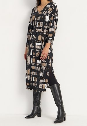 Kobieta nosząca midi sukienkę w czarno-beżowo-biały wzór z długimi rękawami, z rozcięciem po boku, czarne legginsy i czarne skórzane kozaki za kolano.