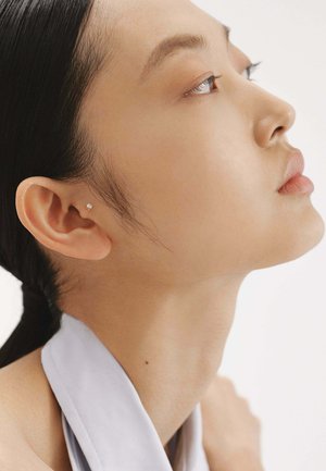 Primer plano de un perfil con piel suave, cabello oscuro recogido, usando un pequeño arete de plata y una tela gris clara drapeada alrededor del cuello.