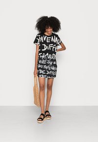 Vestido preto de manga curta com texto branco em destaque, combinado com sandálias pretas de plataforma e uma bolsa de palha beje trançada. Tecido suave, design ajustado.
