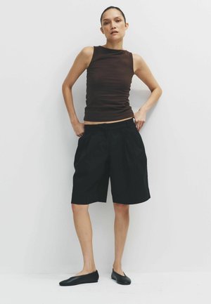 Kobieta stojąca przy białej ścianie, ubrana w bezrękawnik w ciemnobrązowym kolorze, luźne czarne szorty do kolan i czarne buty wsuwane.