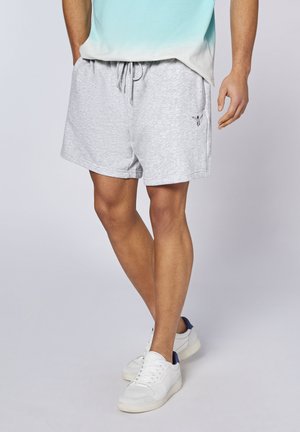 Mann trägt hellgraue Shorts mit Kordelzug, weiße Sneakers und ein türkis-weißes Ombre-Shirt, steht mit gekreuzten Beinen.