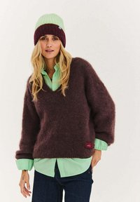 Purpurový chlupatý svetr s výstřihem do V, vrstvený přes světle zelenou košili na knoflíky. Pletená dvoubarevná čepice ve světle zelené a tmavě červené barvě.