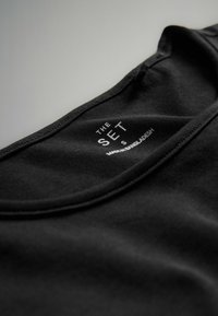 Schwarzes Baumwoll-T-Shirt mit Rundhalsausschnitt, versehen mit einem Etikett, das "THE SET" und "MADE IN BANGLADESH" in weißer Schrift enthält.
