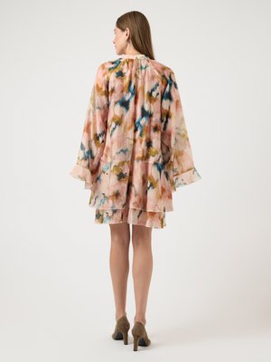 Kvinde med langt hår iført en løs, lagdelt kjole med blomstermønster i rosa, blå og brune nuancer, parret med brune ruskindshæle.